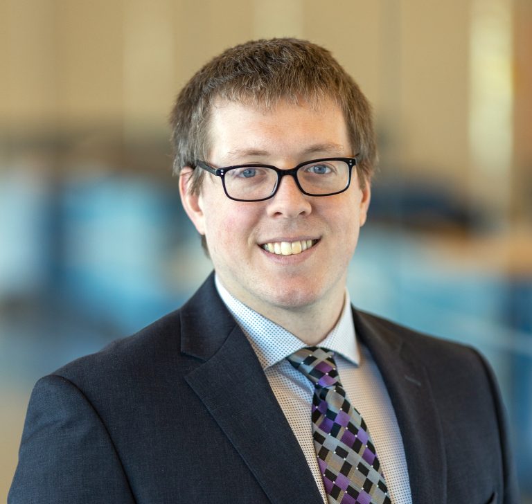 Smiling man wearing glasses, a dark suit, patterned shirt, and purple geometric tie