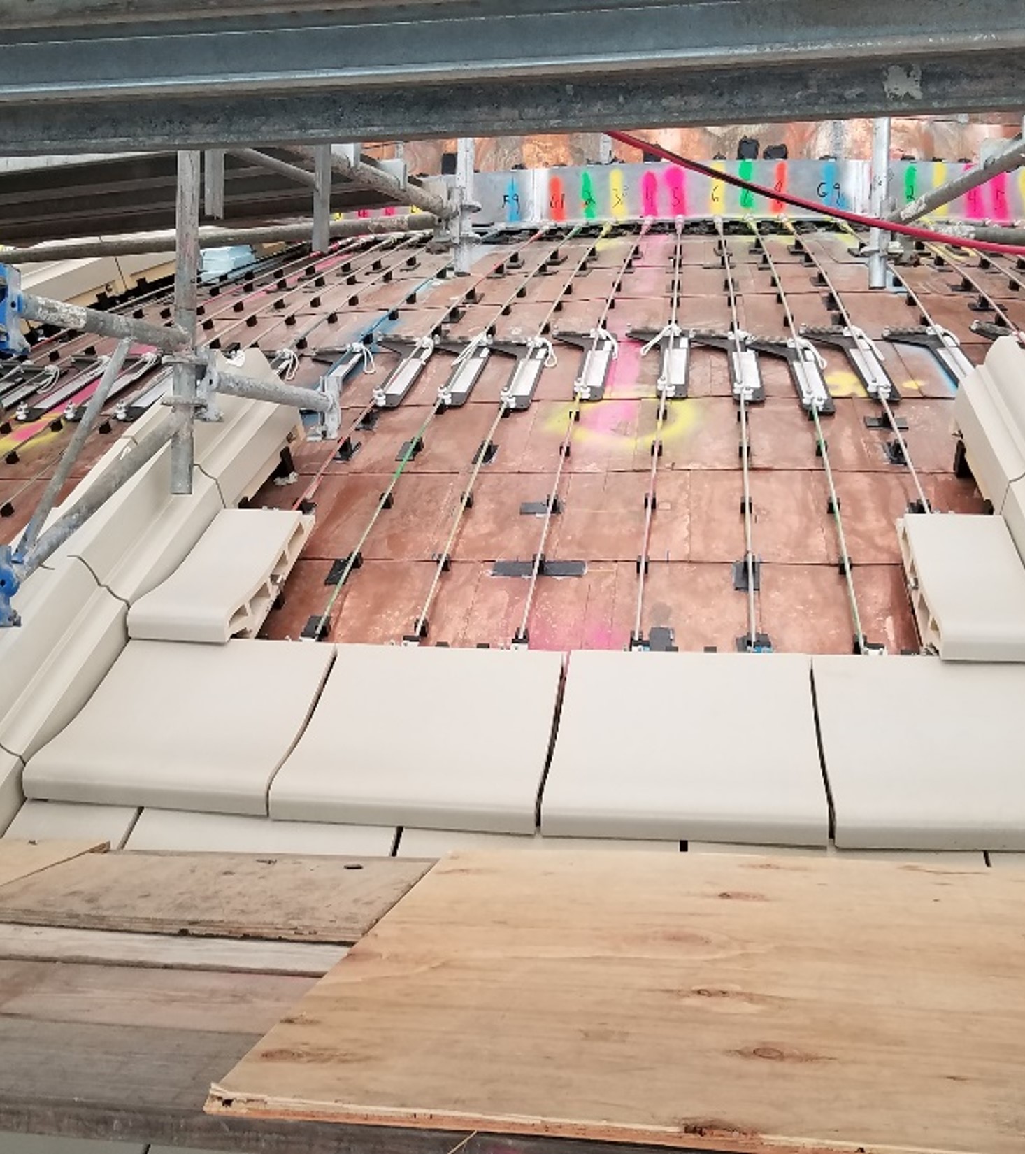 Construction view showing rows of cables and metal fixtures beneath a structure, with scaffolding and color-coded labels