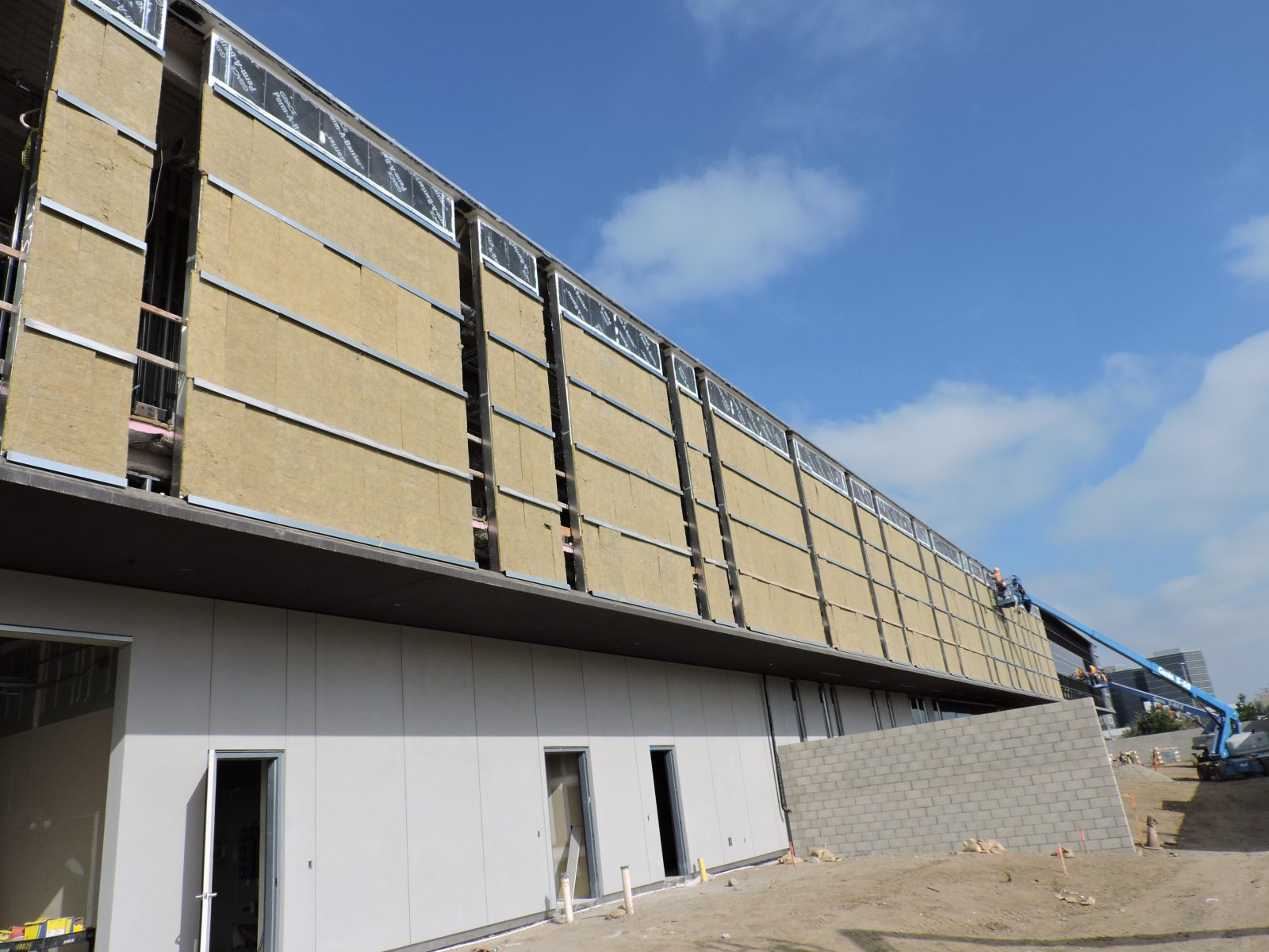 Partially constructed multi-story building with exposed insulation, workers, and construction equipment under a blue sky