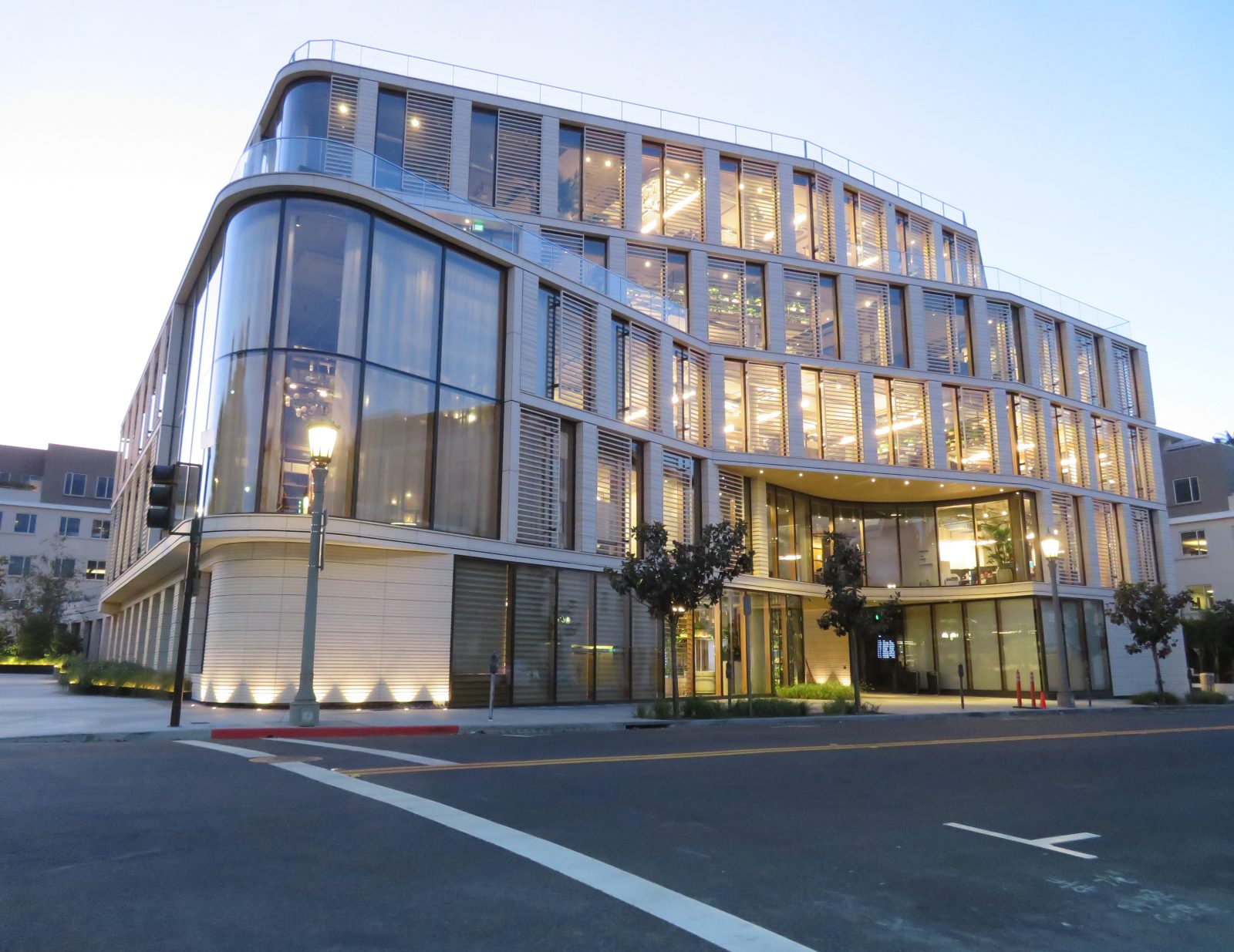 Modern multi-story building with curved glass walls and illuminated windows beside a city street at dusk