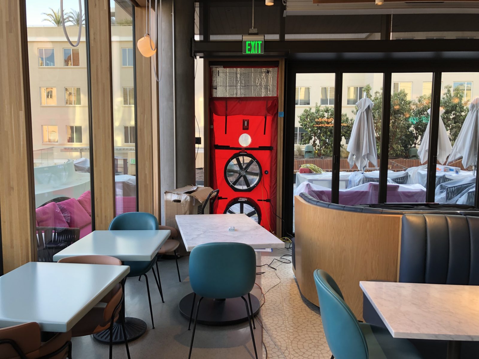 Restaurant dining area with tables and chairs, glass doors, patio umbrellas, and a green EXIT sign above a red fan unit