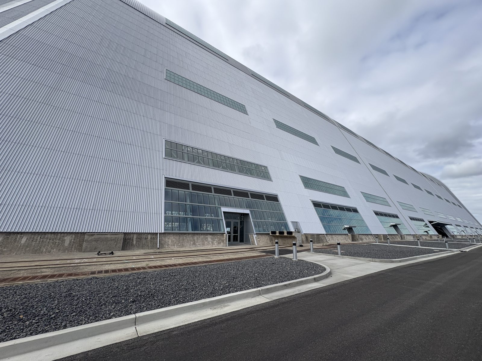 Exterior of a large corrugated-metal industrial building with rows of windows under a cloudy sky