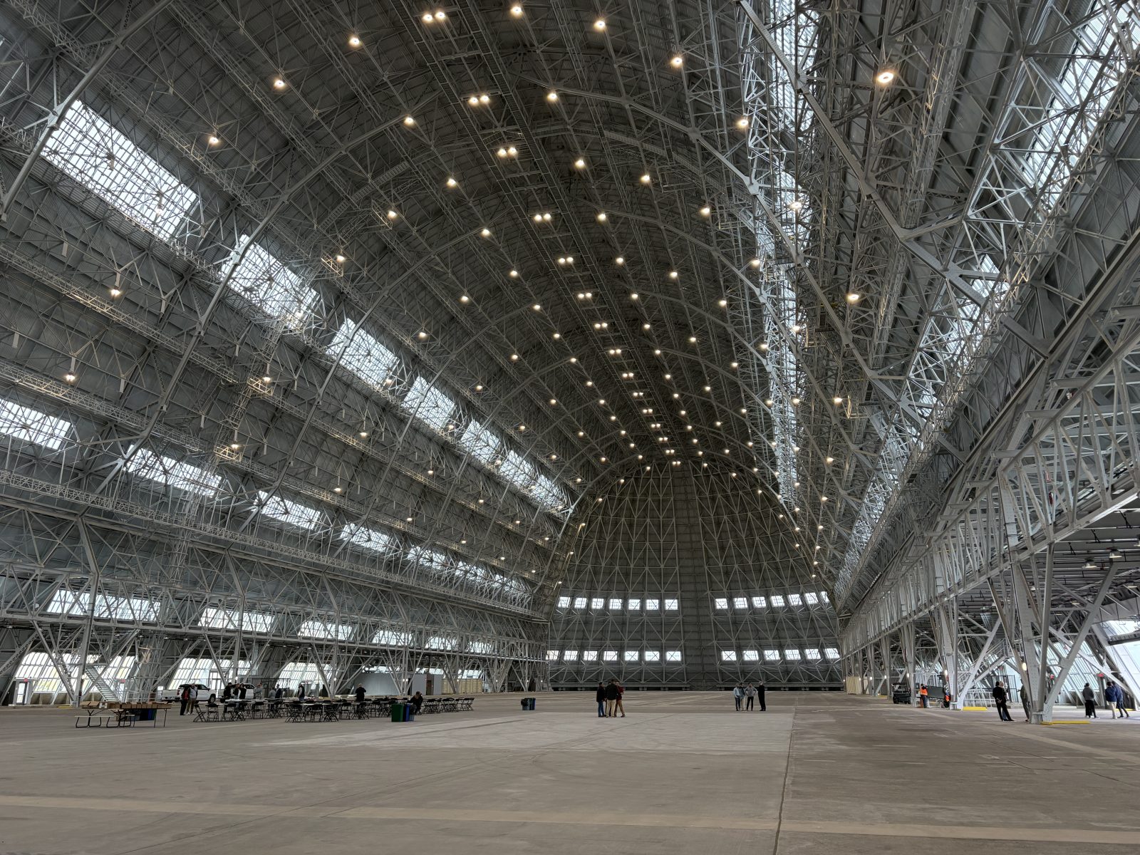 Vast indoor exhibition hall with steel trusses, skylights, rows of lights, tables, and people on the concrete floor