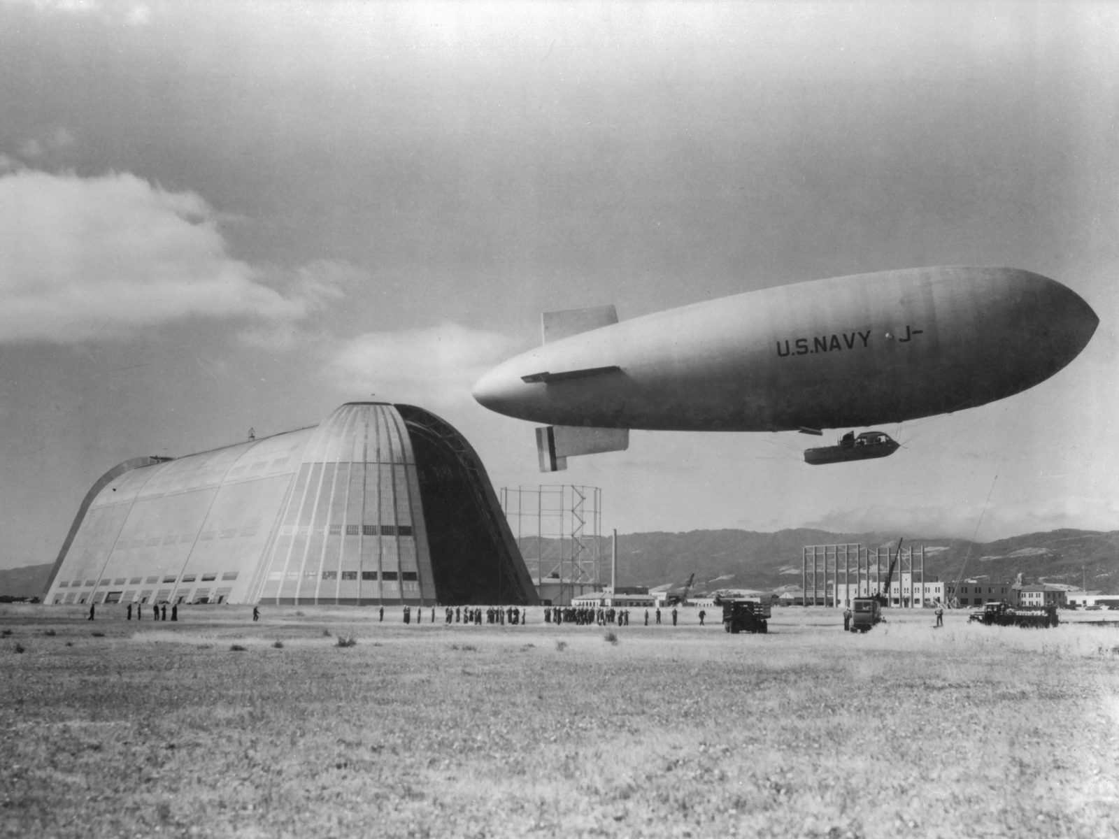U.S. Navy airship marked “J-1” flies over a large hangar as people gather below