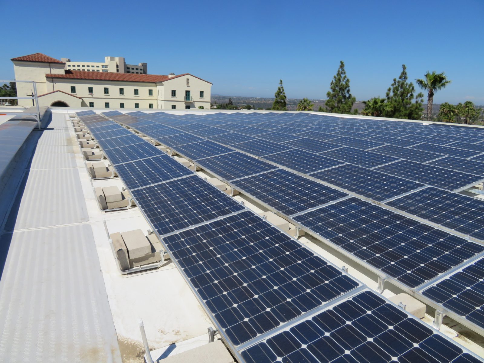 Rows of solar panels cover a rooftop, with a Spanish-style building, palm trees, and a distant landscape under a clear blue sky