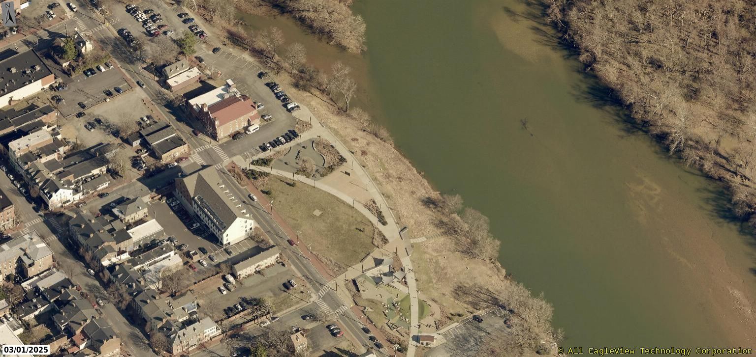 Aerial view of a town bordering a wide river, with buildings, roads, parking lots, and a riverside park