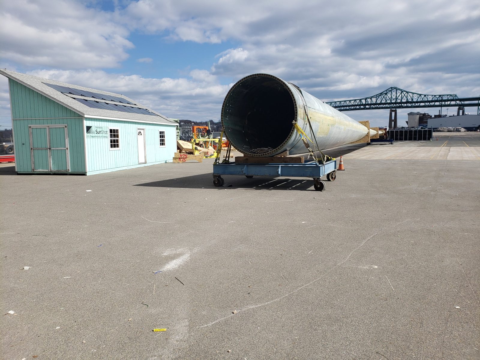 Large cylindrical metal structure secured to a trailer at an industrial site, with a bridge in the background