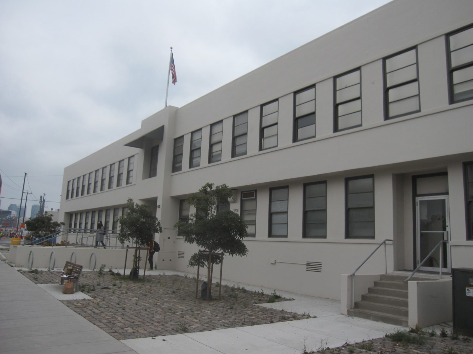 White modern building with rows of windows, entrance steps, trees, and an American flag