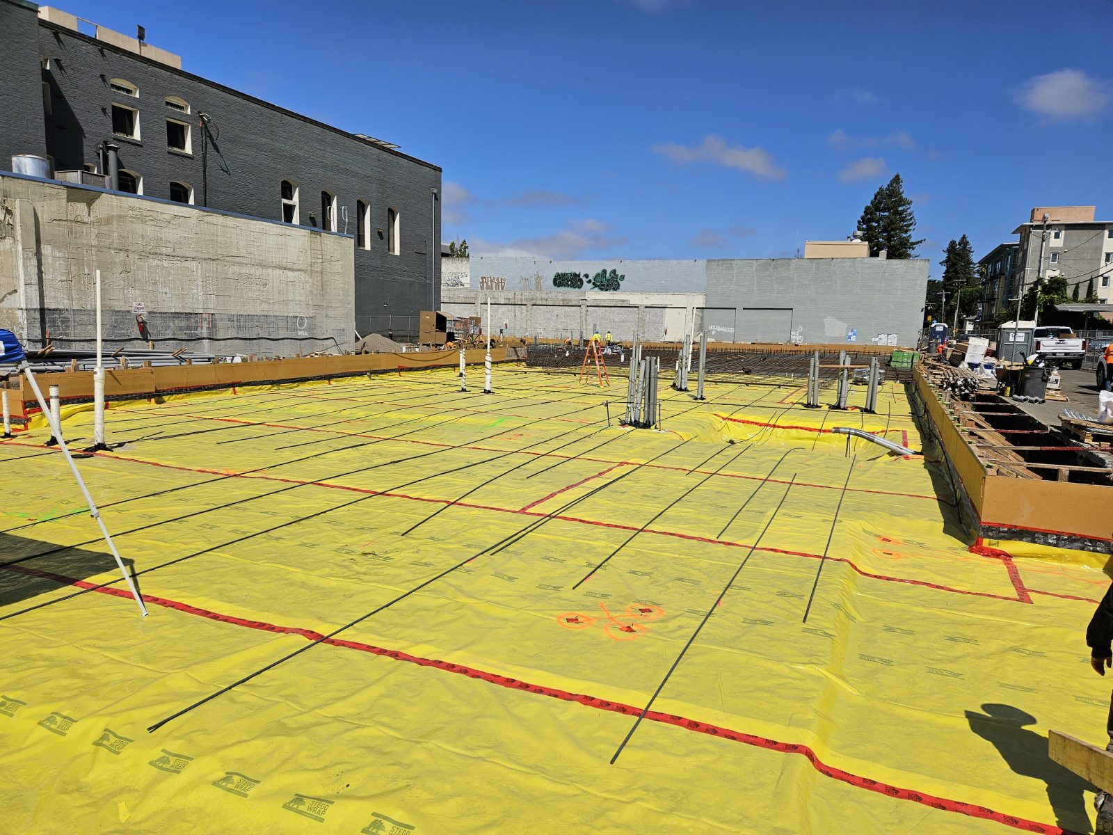 Construction site with yellow vapor barrier, exposed rebar, workers, and surrounding urban buildings