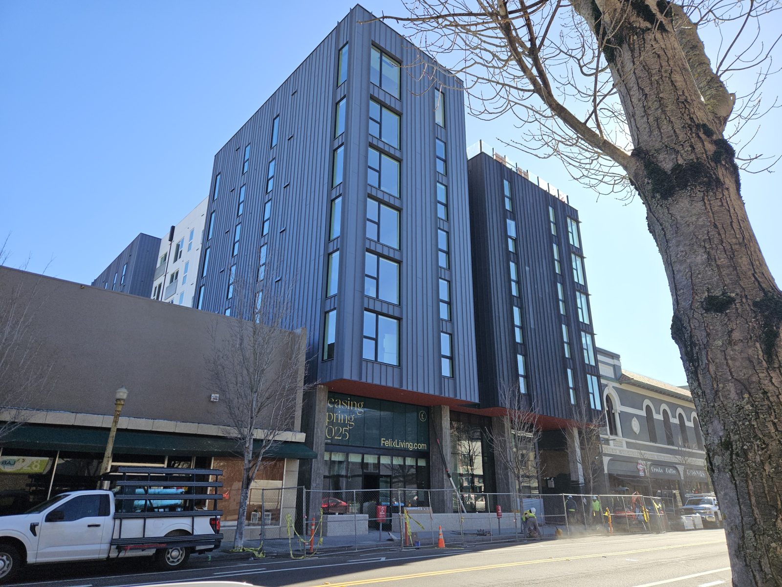 Modern gray multistory building under construction on a city street, with a large tree in the foreground