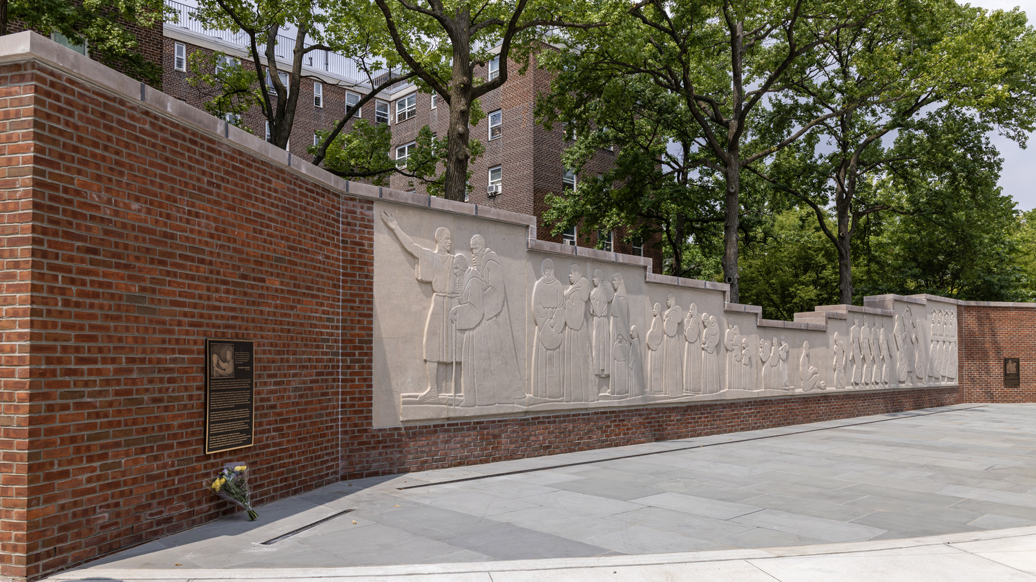 Brick memorial wall featuring a long stone relief of people in procession, with a plaque and flowers