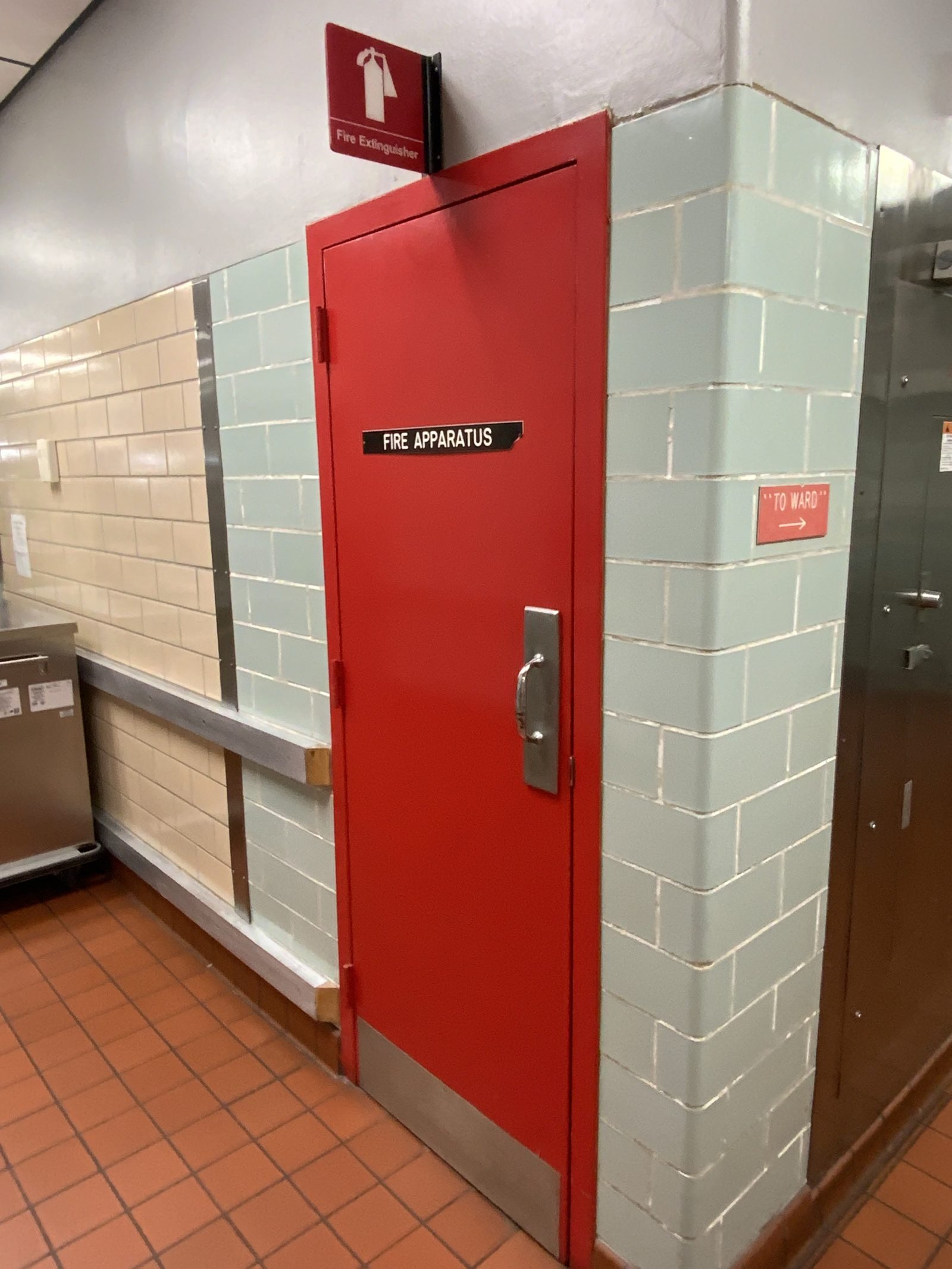 Red fire apparatus cabinet with a fire extinguisher sign in a tiled hallway; nearby sign points to the ward