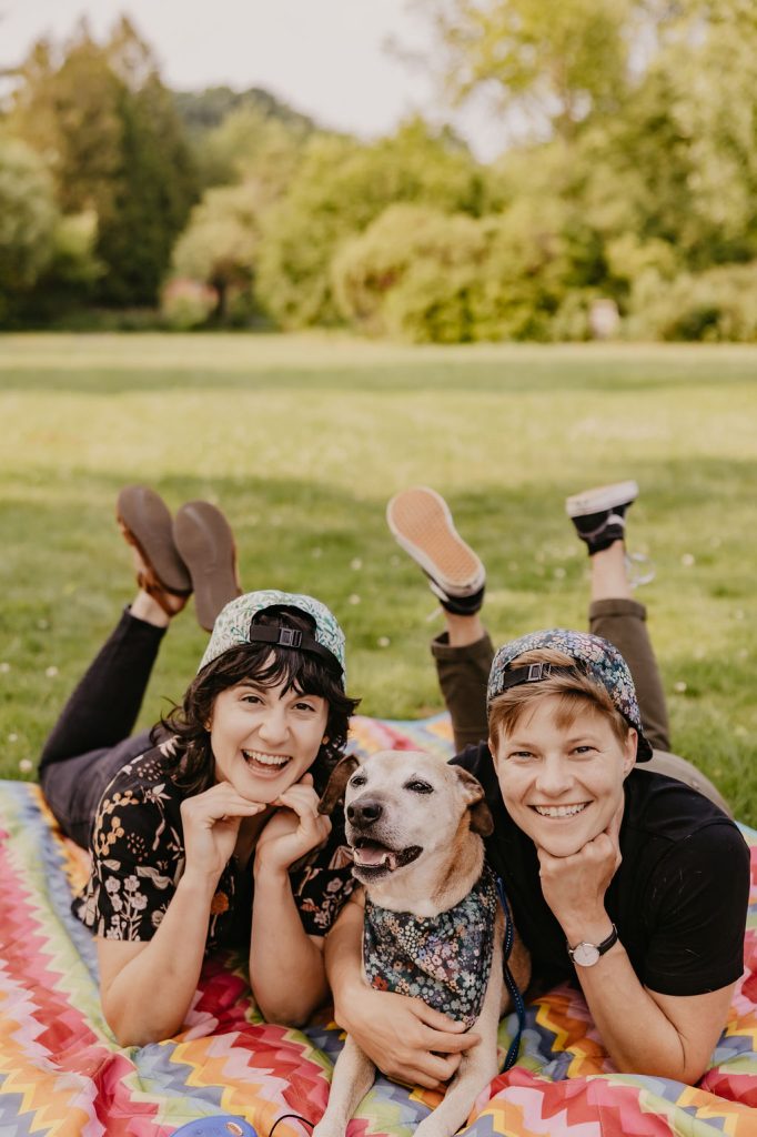 Two people and a dog lying on a colorful blanket in a grassy park, all smiling at the camera. The dog is between them, and both people are wearing patterned caps.