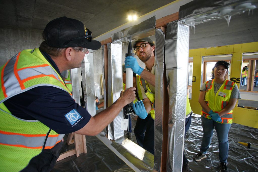 Workers in safety vests install reflective insulation around a structure while a colleague observes