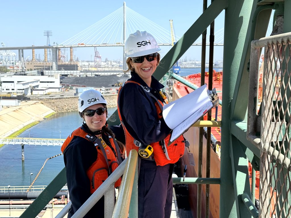 Two workers in safety gear and hard hats stand on an industrial platform near a port, holding a clipboard and smiling at the camera, exemplifying the opportunities available through SGH Careers.