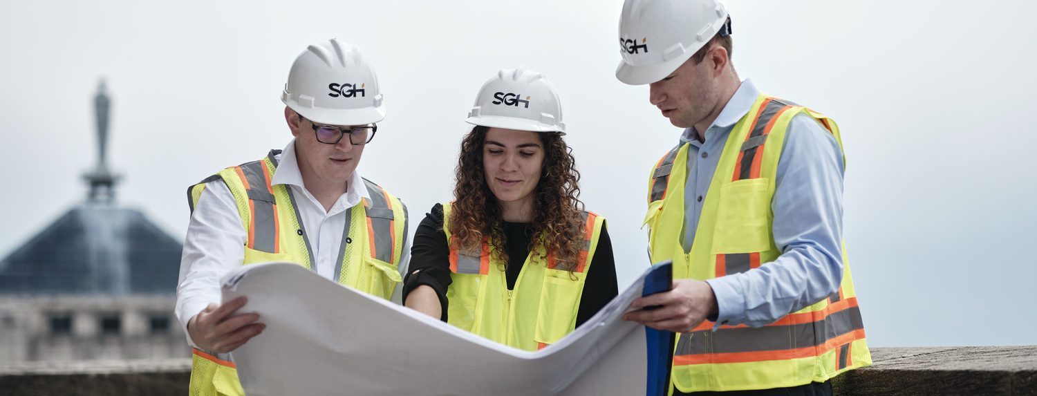Three people in safety vests and hard hats review blueprints outdoors at a construction site, exemplifying the collaborative environment found in SGH Careers.