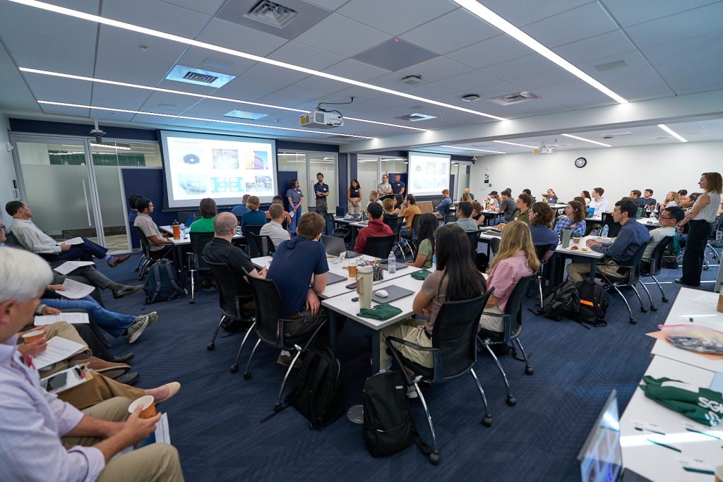 Adults attend a workshop in a classroom while presenters stand beside projected slides