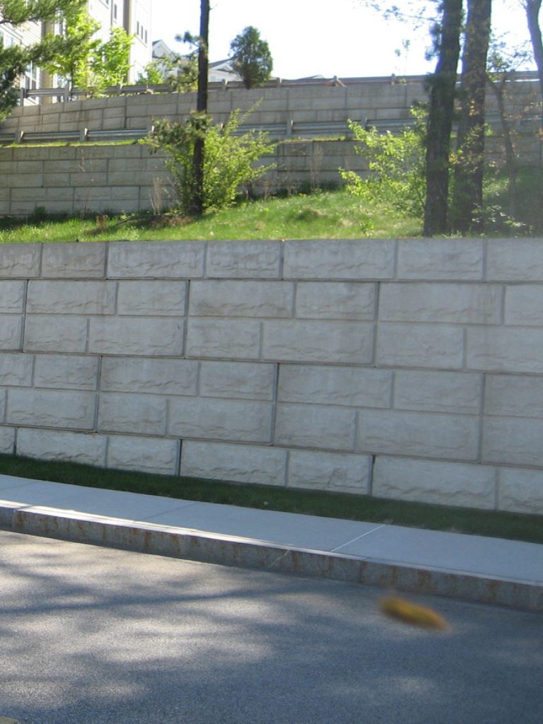 Tiered concrete retaining wall beside a sidewalk, with trees and houses behind it