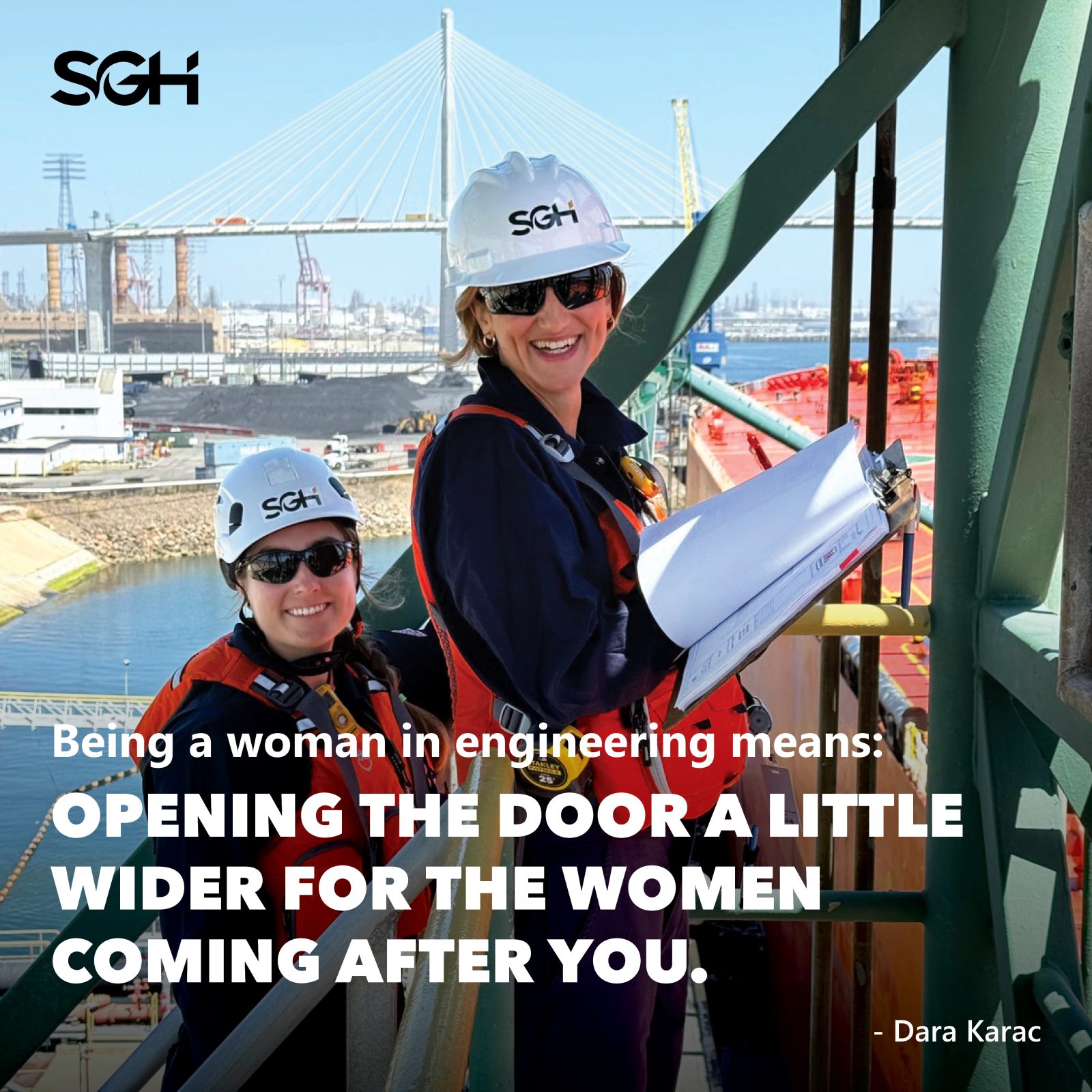 Two women engineers wearing safety gear and helmets stand on a construction site walkway, holding documents, with a bridge and industrial background visible.
