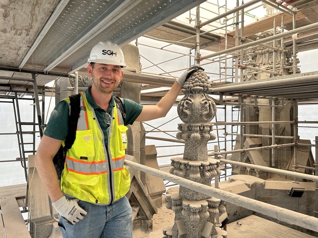 Construction worker in a safety vest stands beside an ornate stone architectural detail amid scaffolding