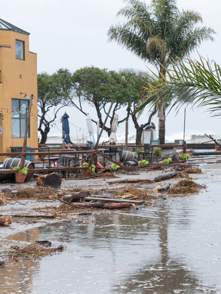 Storm-damaged waterfront restaurant patio covered in debris and standing water, with large waves behind it